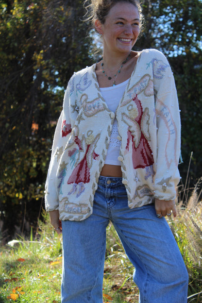 “Soft white cardigan featuring embroidered angel figures and small beaded accents for added texture.”