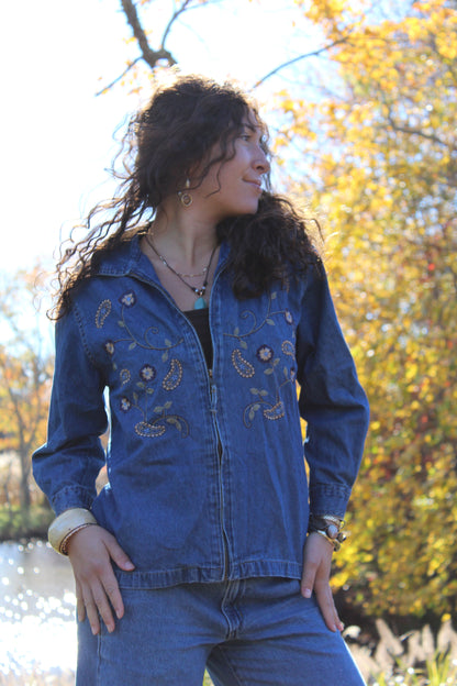 Front view of a medium-wash denim jacket with stitched embroidery and a zipper front.